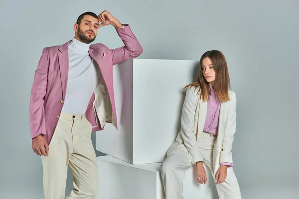 Hombre de moda en blazer lila mirando a la cámara cerca de cubos blancos y mujer elegante sentado en gris - foto de stock