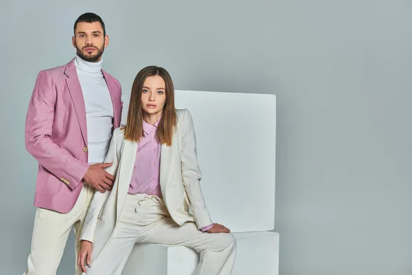 Woman in white suit and man in lilac blazer looking at camera near white cubes on grey, trendy wear — Stock Photo