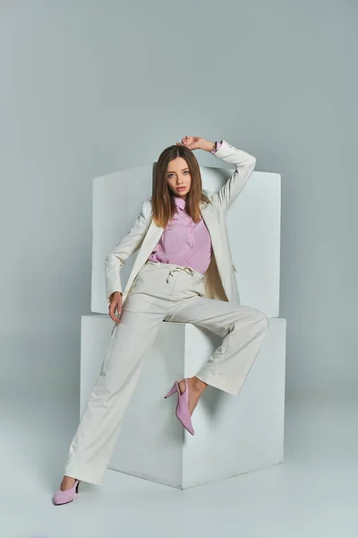 Longitud completa de la mujer atractiva en traje elegante posando y mirando a la cámara cerca de cubos blancos en gris - foto de stock