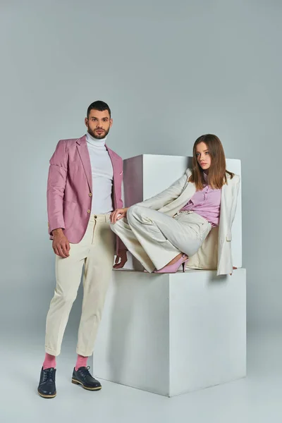 Fashionable man in lilac blazer looking at camera near woman in white suit posing on cubes on grey — Stock Photo