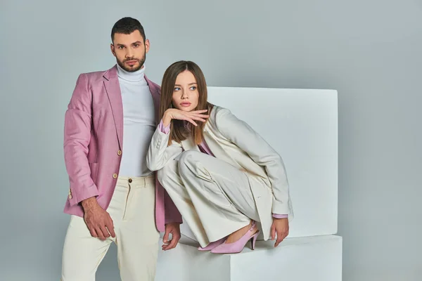 Selbstbewusster Mann in fliederfarbenem Blazer blickt in die Kamera neben stilvoller Frau, die auf weißen Würfeln auf grau posiert — Stockfoto