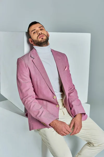 Homme charismatique en blazer lilas à la mode assis sur cube blanc et regardant la caméra sur gris — Photo de stock