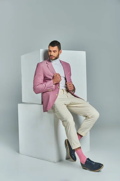 Full length of trendy man in pastel purple blazer and white pants posing near cubes on grey backdrop — Stock Photo