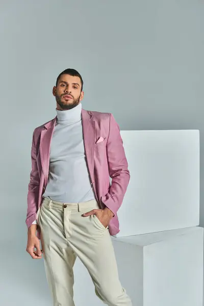 Hombre seguro en chaqueta lila posando con la mano en el bolsillo cerca de cubos blancos en gris, moda de negocios - foto de stock