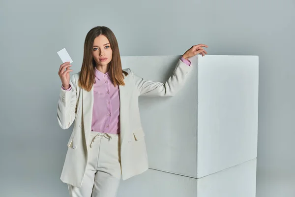 Молода успішна жінка в стильному формальному вбранні, що тримає порожню візитку біля білих кубиків на сірому — стокове фото
