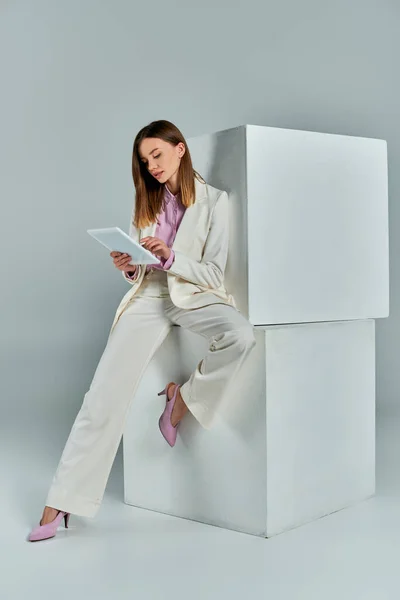 Full length of stylish woman in white suit networking on smartphone near white cubes on grey — Stock Photo