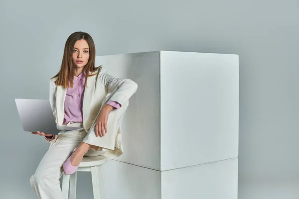 Mujer elegante en traje blanco sentado en el taburete con el ordenador portátil y mirando a la cámara cerca de cubos en gris - foto de stock