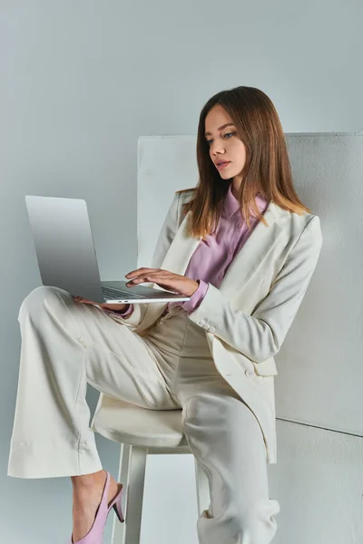Mujer joven en traje elegante en red en el ordenador portátil mientras está sentado en el taburete cerca de cubos blancos en gris - foto de stock