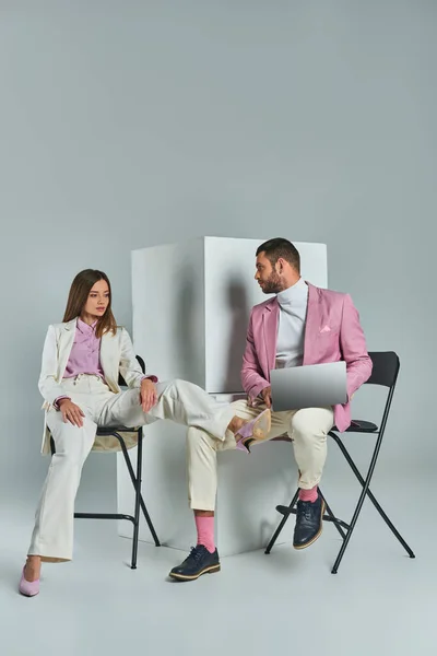Hombre elegante con el ordenador portátil y la mujer joven en traje blanco sentado en sillas cerca de cubos sobre fondo gris - foto de stock