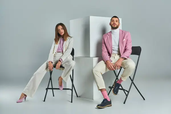 Confident couple in stylish formal wear sitting on chairs near cubes on grey, minimalistic fashion — Stock Photo