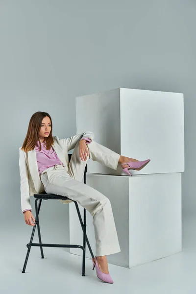 Longitud completa de encantadora mujer de negocios en traje blanco sentado en la silla cerca de cubos blancos en gris - foto de stock