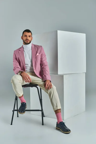 Hombre elegante en traje de negocios sentado en la silla y mirando a la cámara cerca de cubos blancos en gris - foto de stock