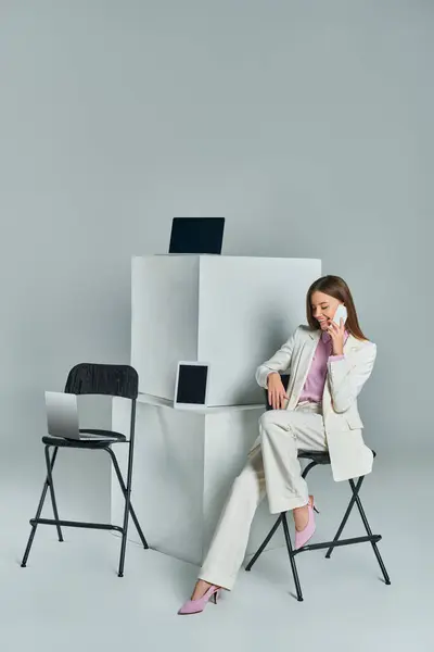 Mujer sonriente sentada en la silla y hablando en el teléfono inteligente cerca de dispositivos en cubos blancos en gris - foto de stock