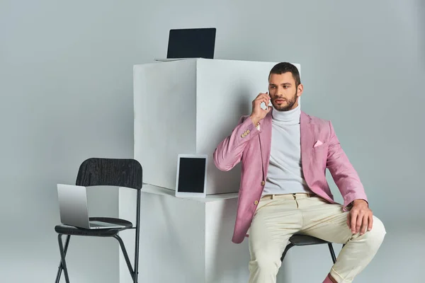 Confident stylish man talking on smartphone while sitting near devices on white cubes on grey — Stock Photo