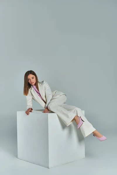 Longitud completa de la mujer joven en traje elegante posando en cubo blanco sobre fondo gris en el estudio - foto de stock