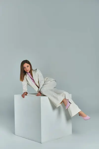 Full length of young woman in elegant suit posing on white cube on grey backdrop in studio — Stock Photo