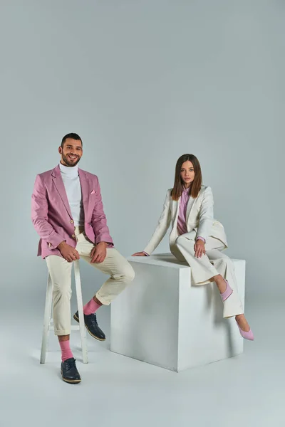 Homem elegante sorrindo na cadeira perto da mulher de terno posando no cubo branco no cinza, moda de negócios — Fotografia de Stock