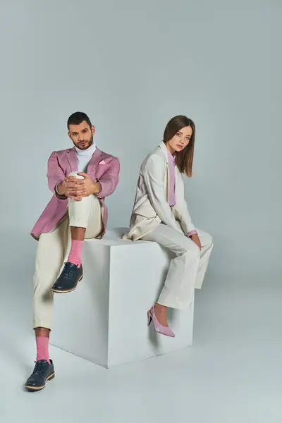Модная деловая пара в элегантной формальной одежде сидит на белом кубе и смотрит в камеру на сером — стоковое фото