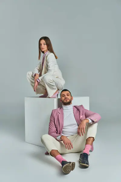 Homme en blazer lilas assis sur le sol près de la femme en costume élégant posant sur cube blanc sur gris — Photo de stock