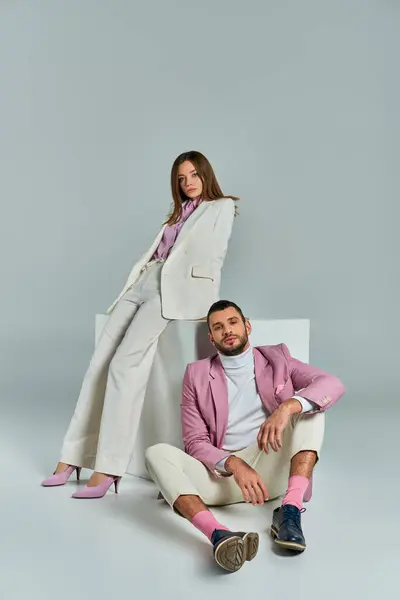 Man in lilac blazer sitting on floor near woman in stylish suit posing near white cube on grey — Stock Photo