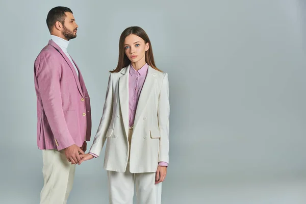 Jeune femme en costume blanc tenant la main avec l'homme en blazer lilas et regardant la caméra sur gris — Photo de stock