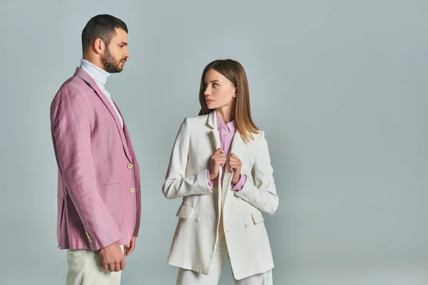 Femme élégante en costume blanc regardant l'homme confiant en blazer lilas sur gris, couple d'affaires moderne — Photo de stock