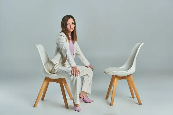 Junge und stilvolle Frau im weißen Anzug sitzt auf einem Sessel und blickt in die Kamera vor grauem Hintergrund — Stockfoto