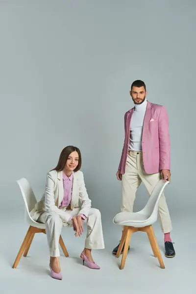 Femme heureuse en costume blanc assis dans un fauteuil près de l'homme en blazer lilas regardant la caméra sur gris — Photo de stock