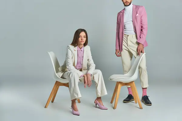 Young woman in white elegant suit sitting in armchair and looking at camera near stylish man on grey — Stock Photo