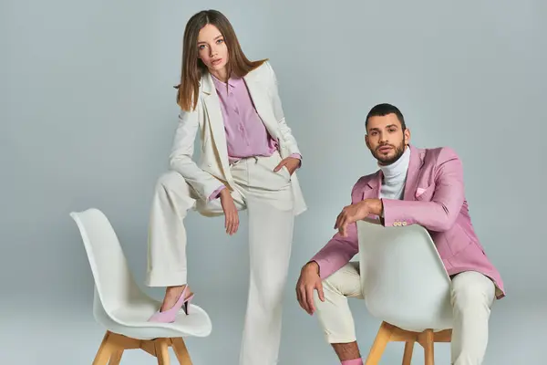 Trendy man in lilac blazer sitting near woman in white elegant suit posing with armchair on grey — Stock Photo
