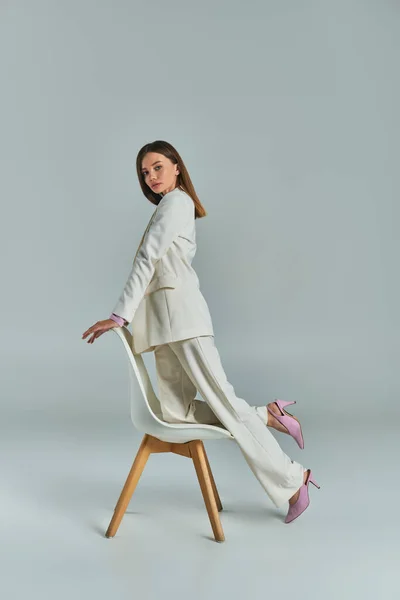 Modern woman in white elegant suit looking at camera and posing with armchair on grey, full length — Stock Photo