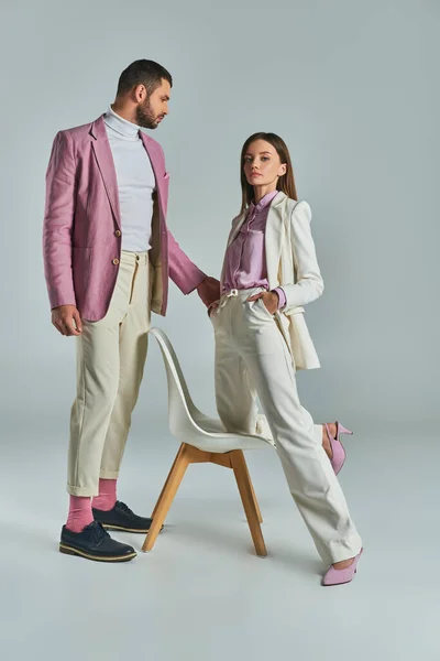 Confident woman in white suit with hands in pockets near man in lilac blazer and armchair on grey — Stock Photo