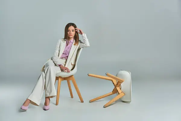Full length of modern woman in white suit sitting on armchair and looking at camera on grey — Stock Photo