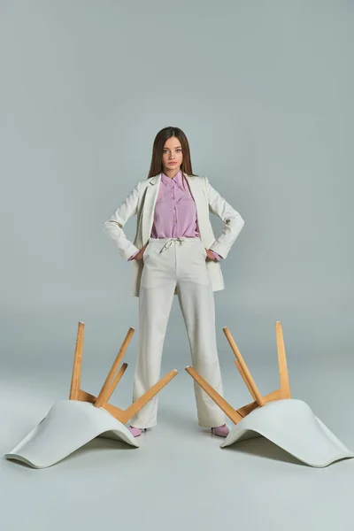Empresaria confiada en traje blanco de pie con las manos en las caderas cerca de sillones volteados en gris - foto de stock