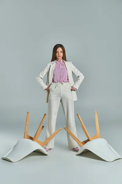 Confident businesswoman in white suit standing with hands on hips near overturned armchairs on grey — Stock Photo