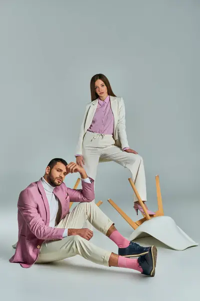 Couple à la mode dans une élégante tenue formelle posant avec des fauteuils renversés sur gris, mode moderne — Photo de stock