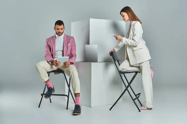 Pareja elegante con teléfono inteligente y tableta digital cerca del ordenador portátil en cubos blancos sobre fondo gris - foto de stock