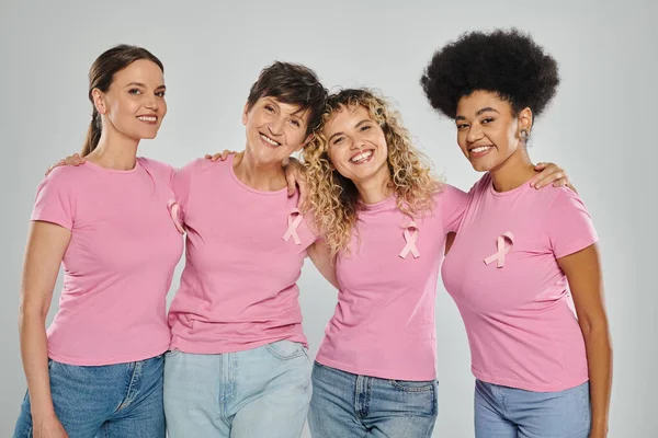 Breast cancer awareness, cheerful interracial women with pink ribbons hugging on grey, diversity — Stock Photo