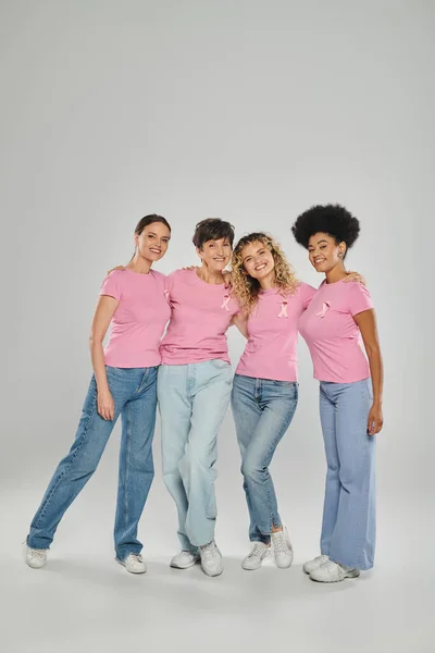 Concienciación sobre el cáncer de mama, alegres mujeres multiculturales con cintas rosas abrazándose al gris, diversidad - foto de stock