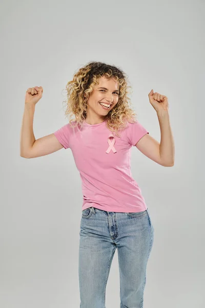 Breast cancer awareness, excited curly woman with pink ribbon looking at camera on grey background — Stock Photo