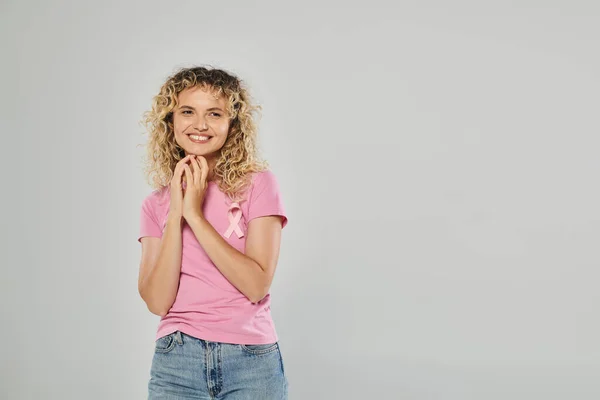Breast cancer awareness, cheerful curly woman with pink ribbon looking at camera on grey background — Stock Photo