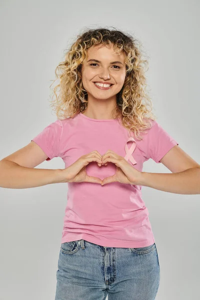 Усвідомлення раку молочної залози, щаслива жінка з рожевою стрічкою, що показує знак серця на сірому фоні, концепція — стокове фото
