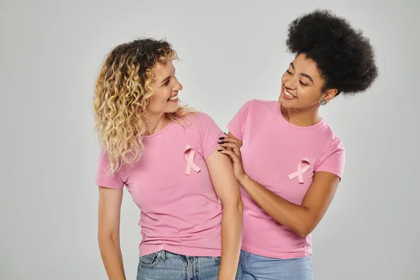 Breast cancer awareness, cheerful multicultural women with pink ribbons on grey backdrop, diversity — Stock Photo