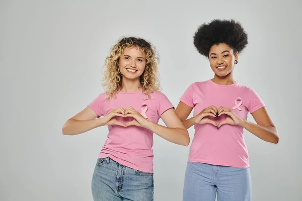 Concienciación sobre el cáncer de mama, mujeres multiculturales felices con cintas rosadas sobre fondo gris, signo cardíaco - foto de stock