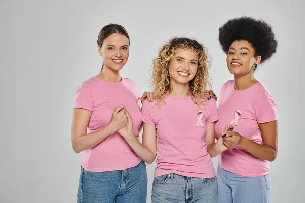 Breast cancer awareness, happy interracial women with pink ribbons holding hands on grey, diversity — Stock Photo