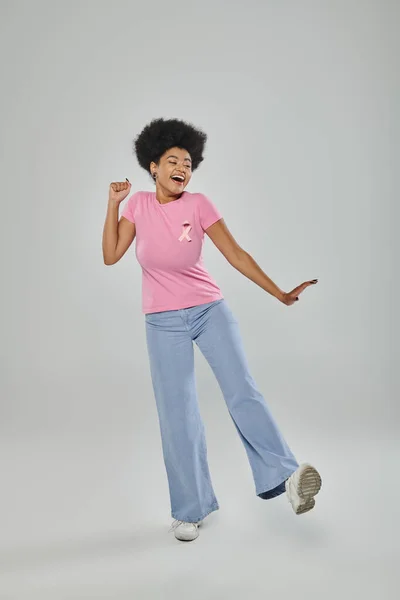Обізнаність про рак молочної залози, схвильована афроамериканська жінка з рожевою стрічкою на сірому, підтримка кампанії — стокове фото