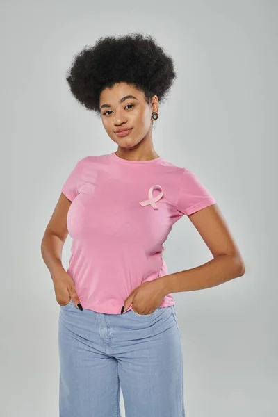 Breast cancer awareness, smile, african american woman with pink ribbon on grey, hands in pockets — Stock Photo