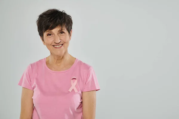Breast cancer awareness, happy middle aged woman with pink ribbon, grey backdrop, portrait — Stock Photo