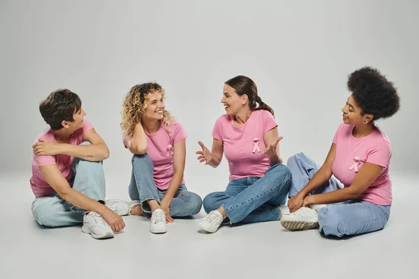 Glückliche interrassische Frauen unterschiedlichen Alters plaudern und sitzen auf grauem Hintergrund, Brustkrebs Bewusstsein — Stockfoto