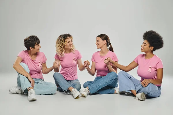 Glückliche interrassische Frauen unterschiedlichen Alters, die Hände vor grauem Hintergrund halten, Bewusstsein für Brustkrebs — Stockfoto